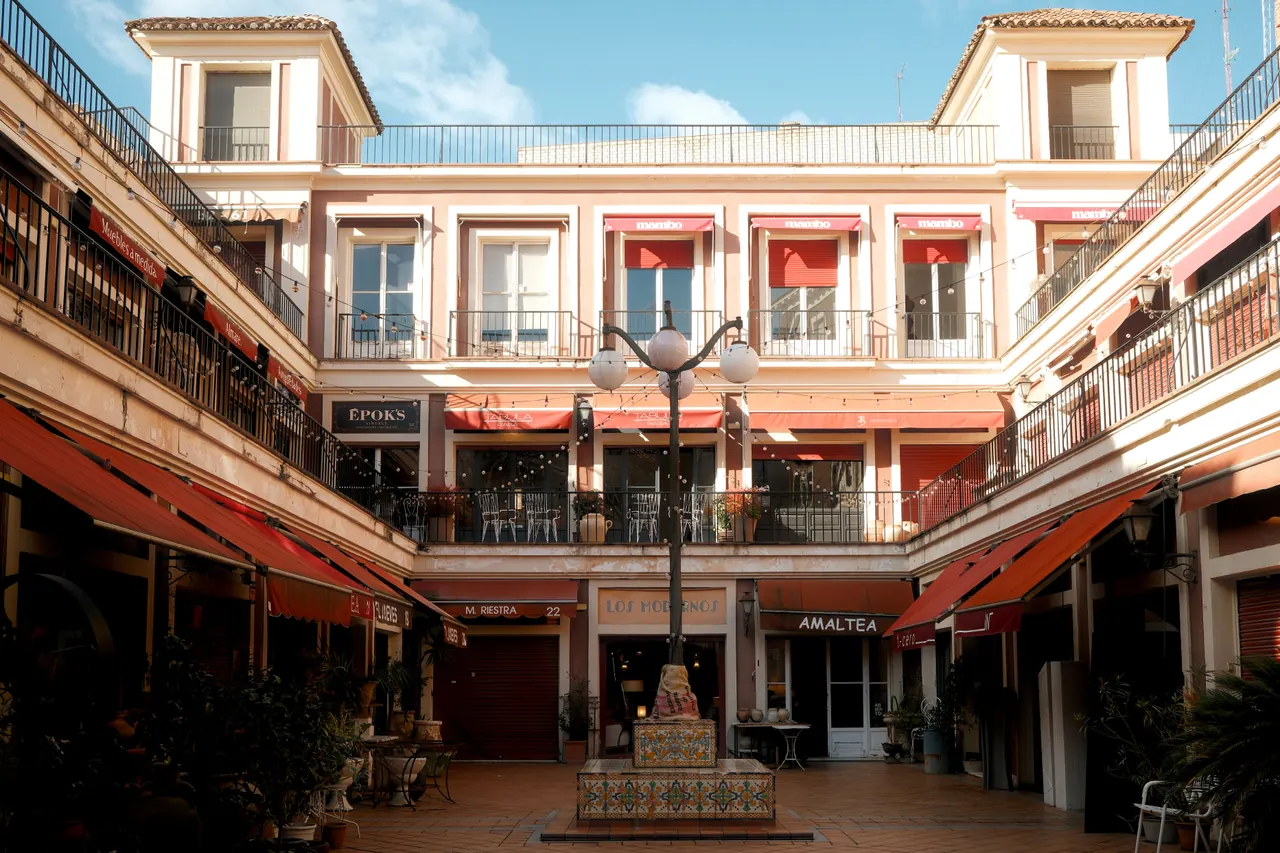 Cour intérieure bordée de boutiques aux stores rouges, autour d'une fontaine en céramique.