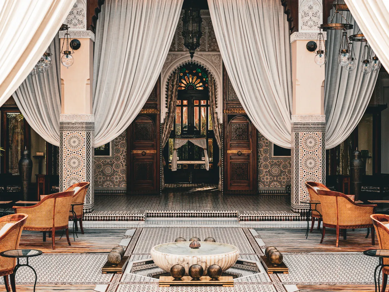 Patio de riad : fontaine en marbre, zelliges, voilages blancs et fauteuils en rotin.
